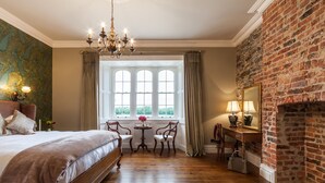 A bedroom with a large bed, a desk, a chair, a small table with a vase, and a fireplace.