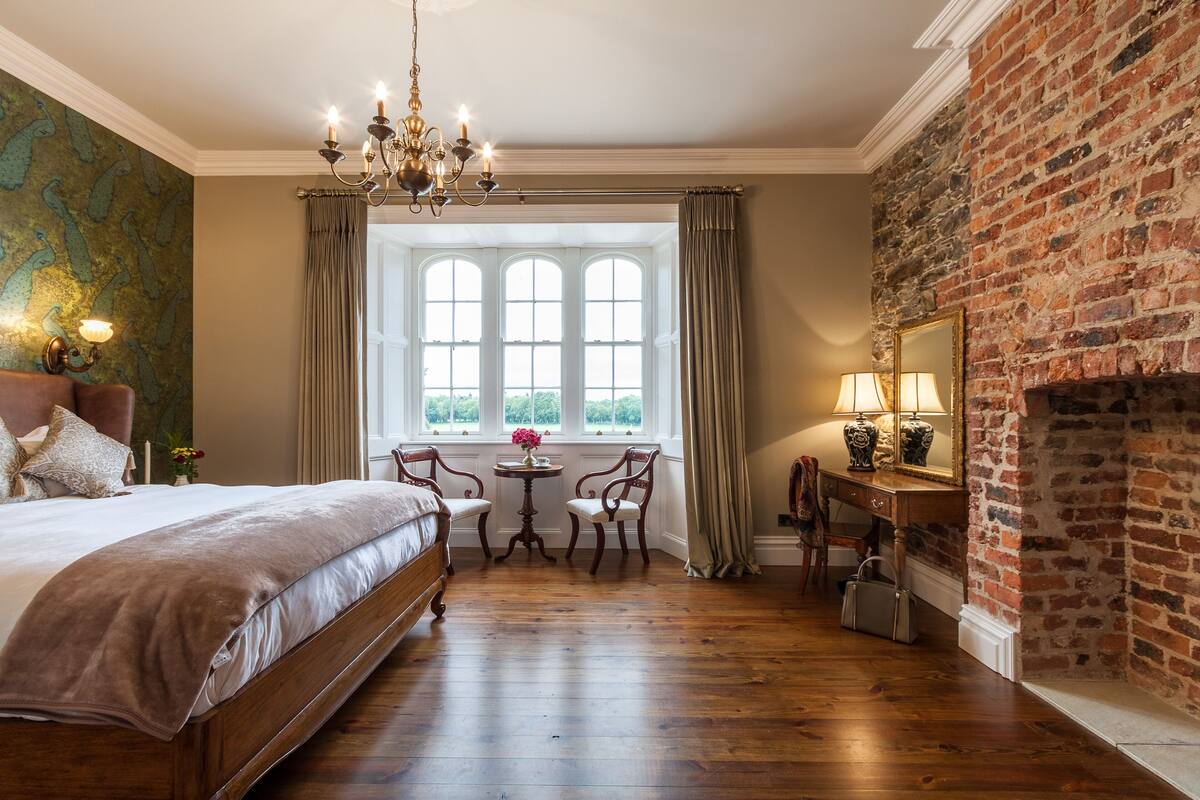 Un dormitorio con cama, escritorio, silla, chimenea y ventana grande.