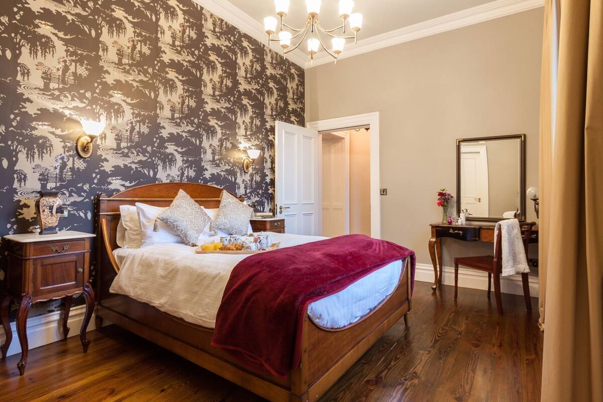 Un dormitorio con una cama de madera, una mesita de noche, un aparador, un espejo y una puerta que da a otra habitación.