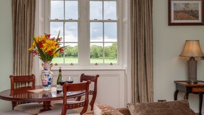 A dining area with a table, chairs, a vase with flowers, a bottle of wine, and a view of greenery through large windows.