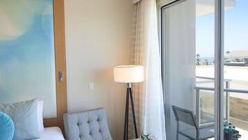 Habitación de hotel con una cama, un sillón gris, una lámpara de pie y vista al océano a través de un ventanal grande.