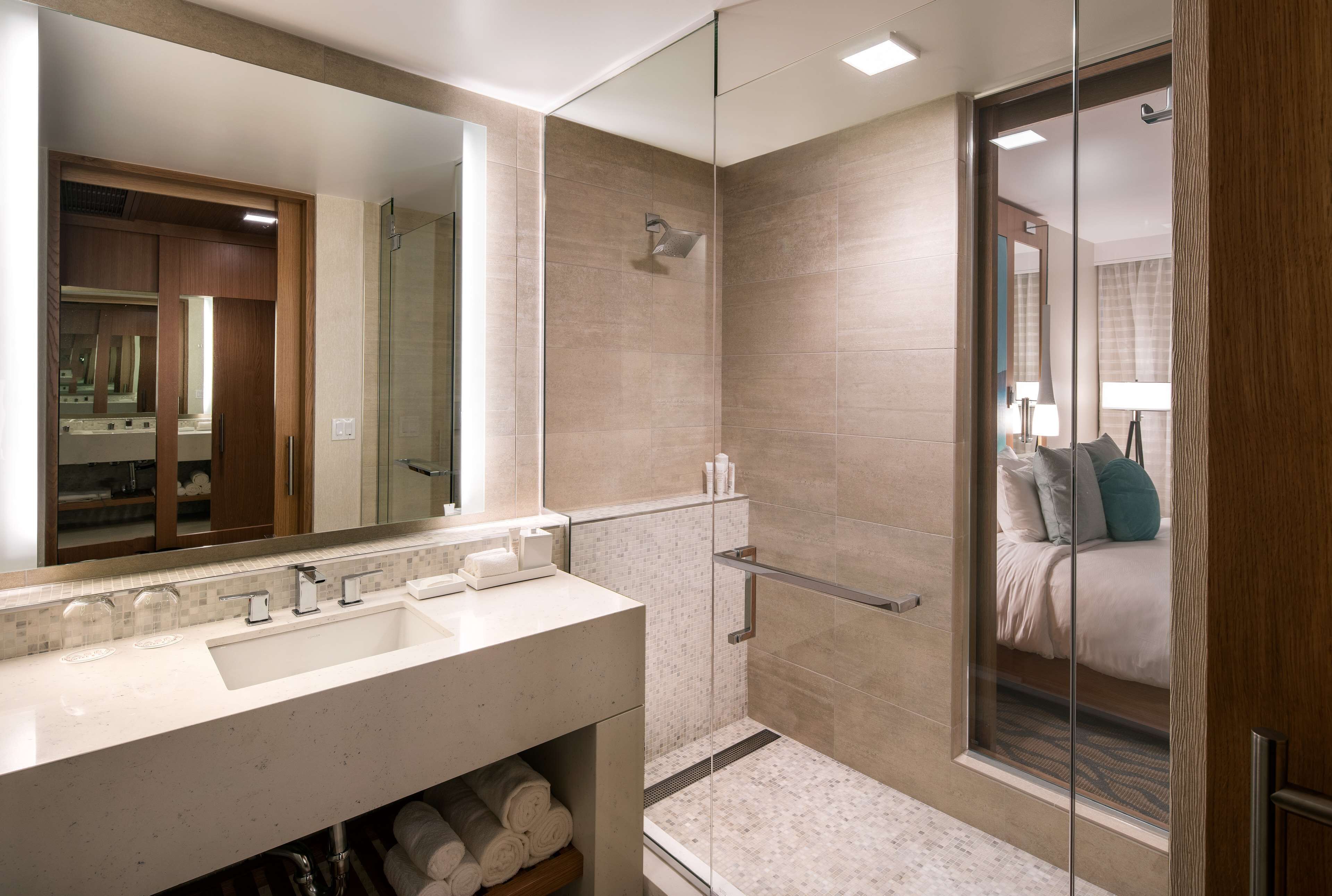Un baño moderno con un espejo grande, cabina de ducha con mampara de vidrio y una cama con almohadas visible a través del cristal.