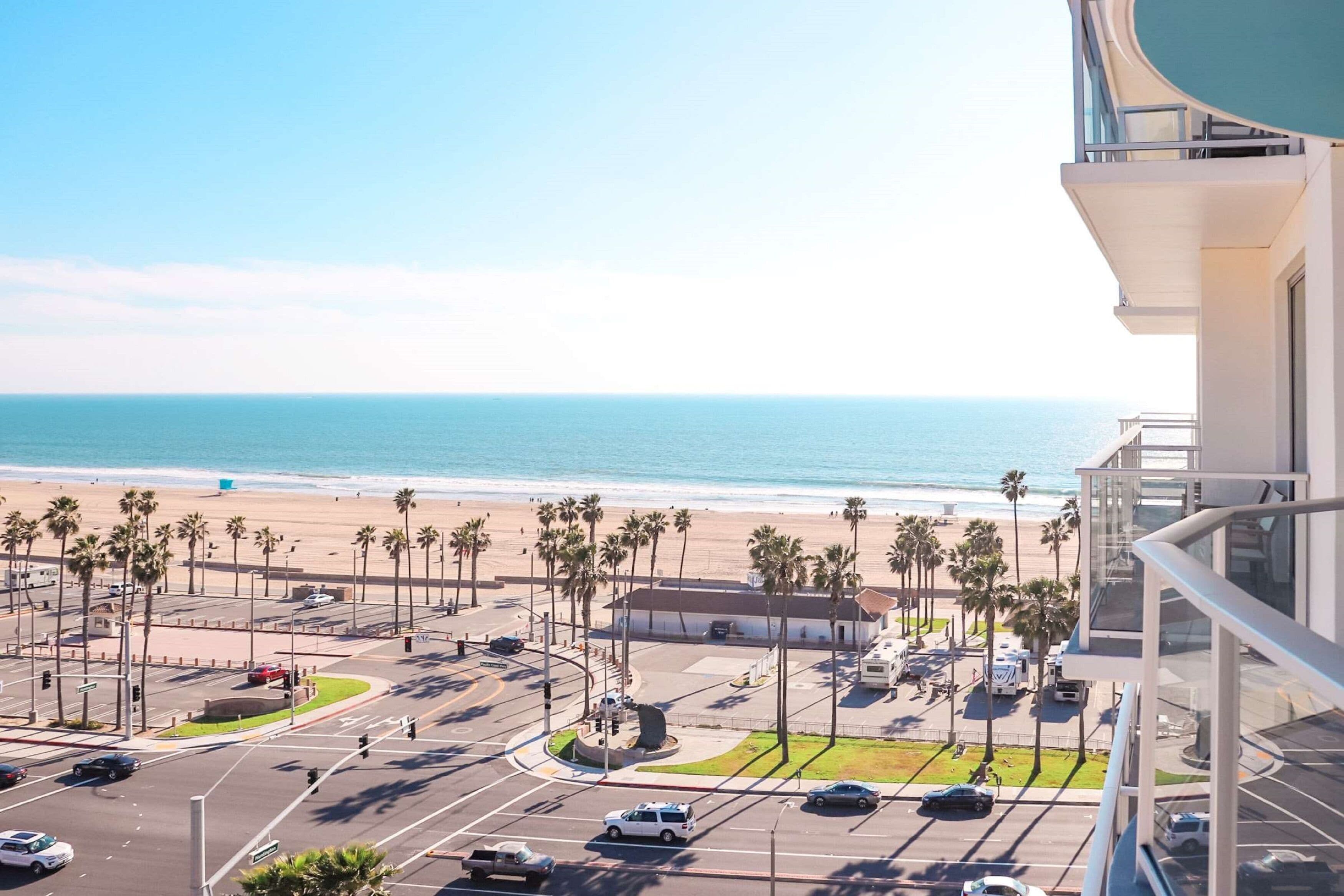 Uitzicht op de kust vanaf een balkon, met uitzicht op een strand, palmbomen, een parkeerplaats en een heldere lucht.