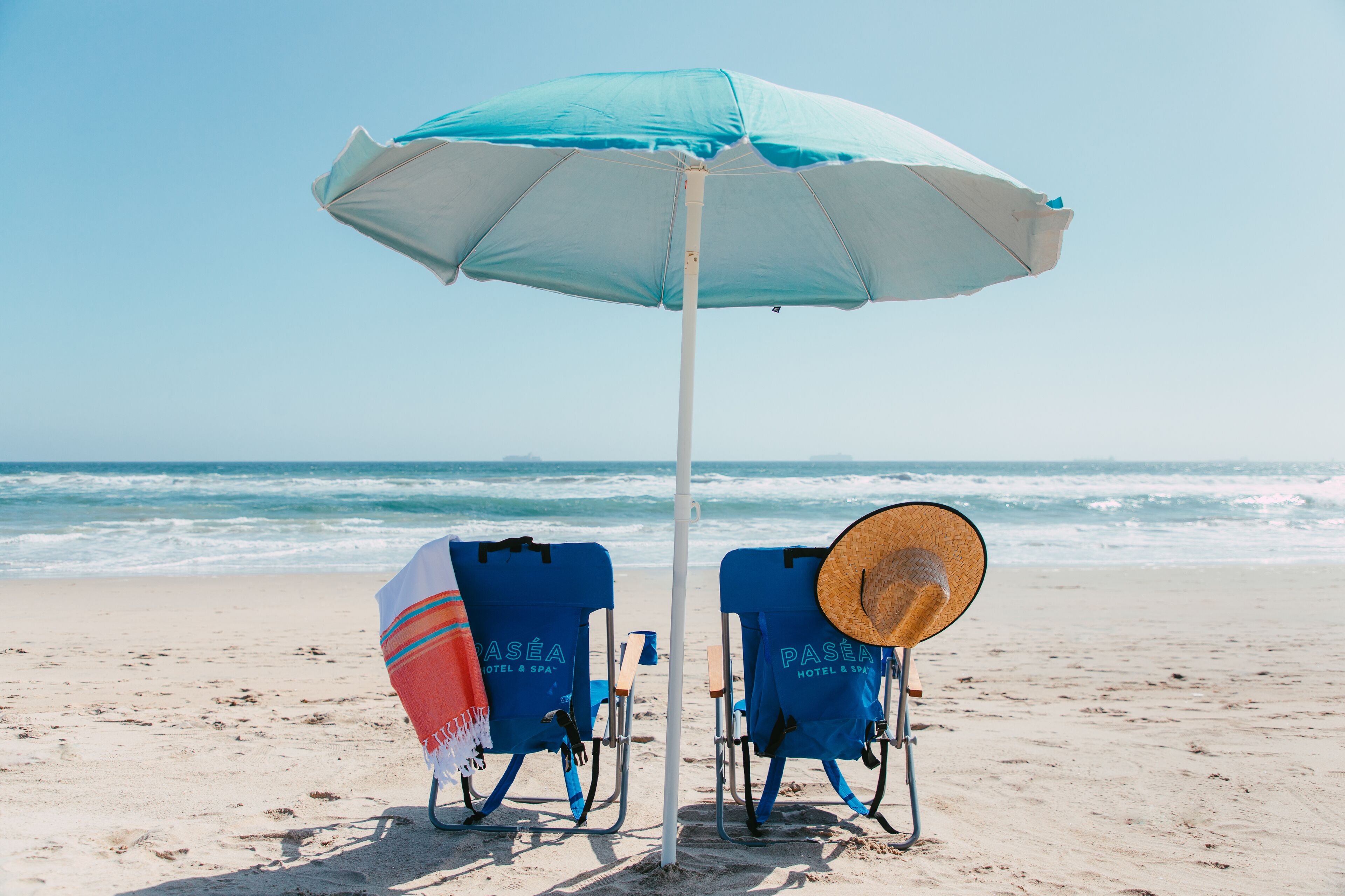 Zwei Strandstühle mit Sonnenschirm und Strohhut im Sand.