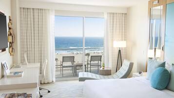 Une chambre d’hôtel avec un grand lit, un bureau avec une chaise, une petite table avec une lampe, et une vue sur la plage et l’océan.