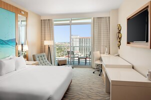 Habitación de hotel con una cama grande, un escritorio, una silla y un balcón con vistas a la ciudad.