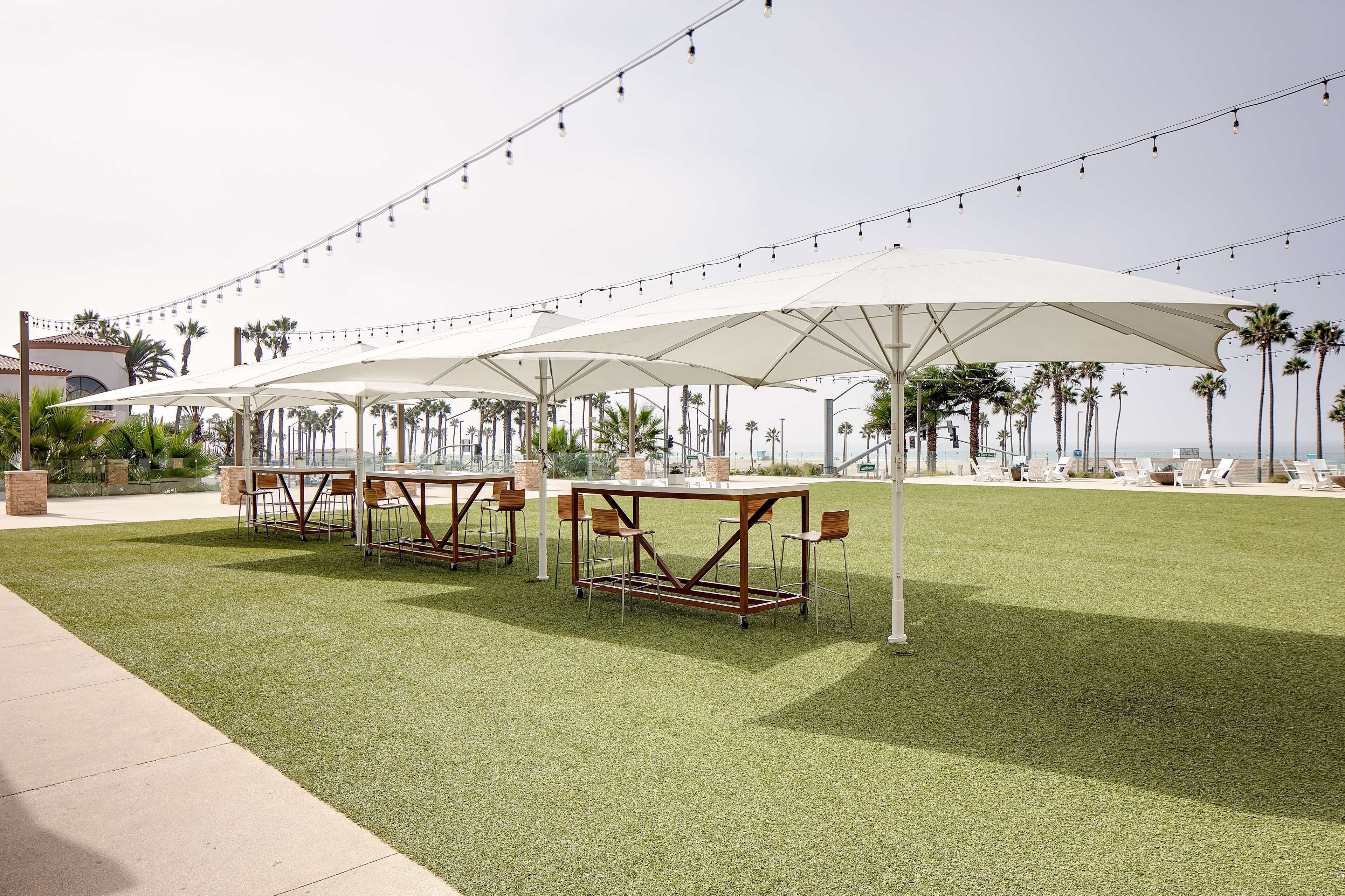 Un espace extérieur aménagé avec des tables et des chaises, protégé par de grands parasols, entouré de palmiers et offrant une vue sur l’océan.