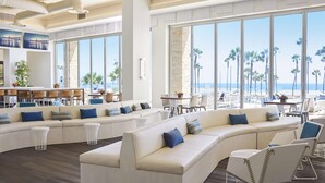 Un salón amplio con un sofá blanco curvo grande, varias sillas y una mesa de comedor. Ventanas amplias ofrecen vistas a la playa y palmeras.