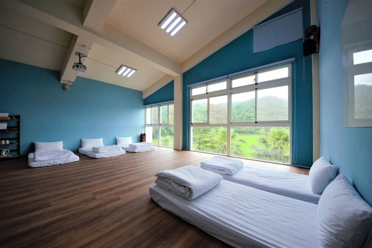 Una habitación amplia con ventanas grandes, piso de madera y camas ordenadas.