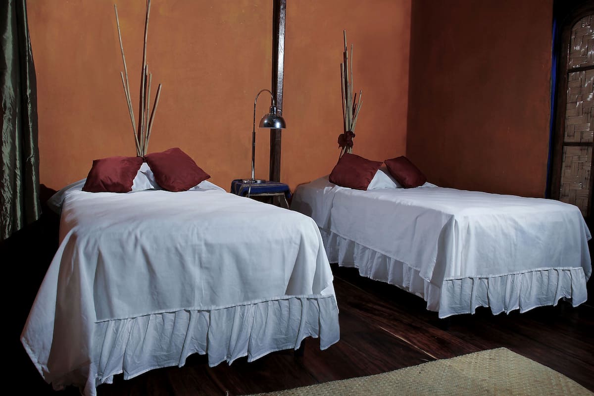 Two beds with white bedding and red pillows, a tall lamp, and a woven basket on a wooden floor.
