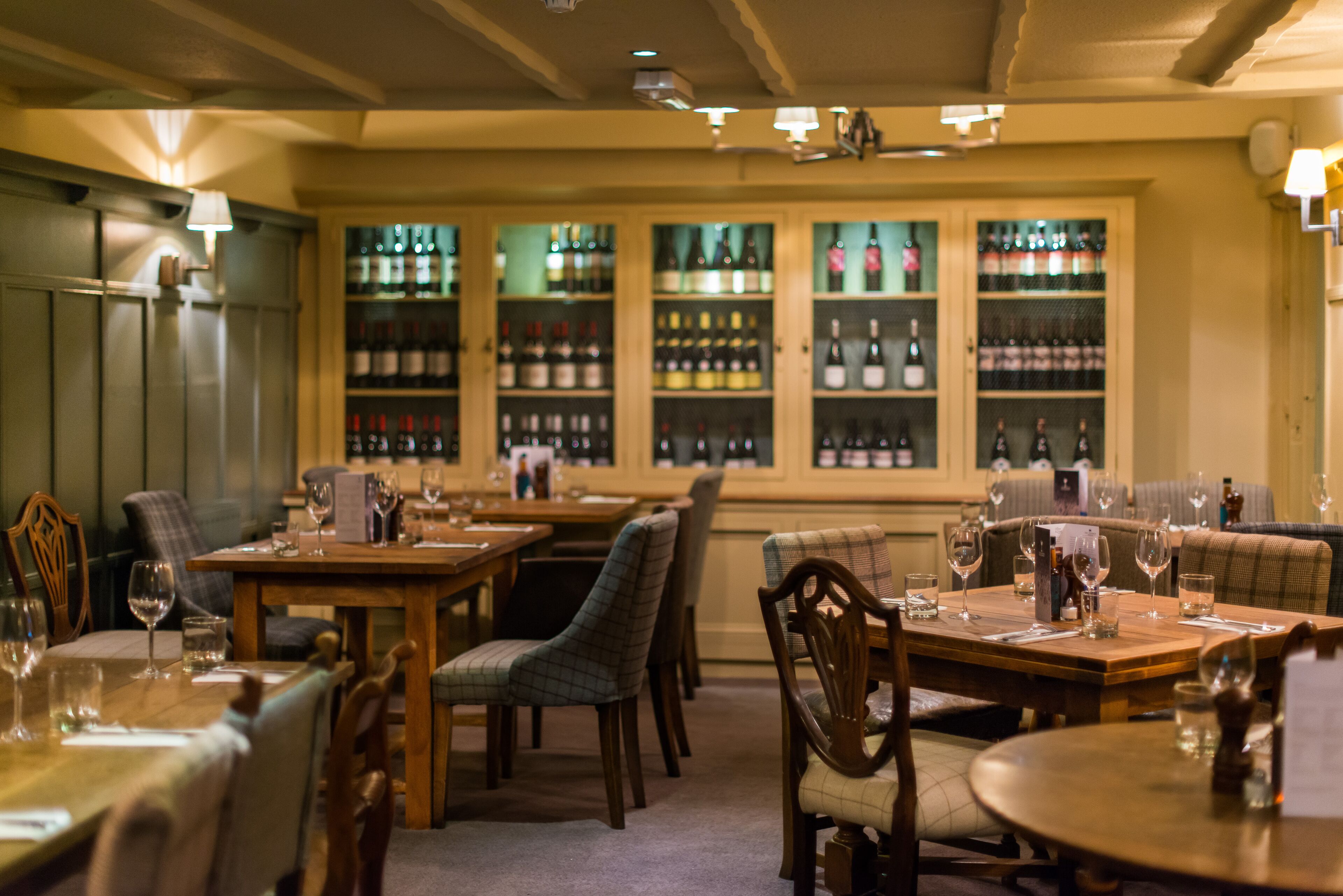 A restaurant with multiple dining tables and chairs, a bar area with shelves of bottles, and a view through large windows.