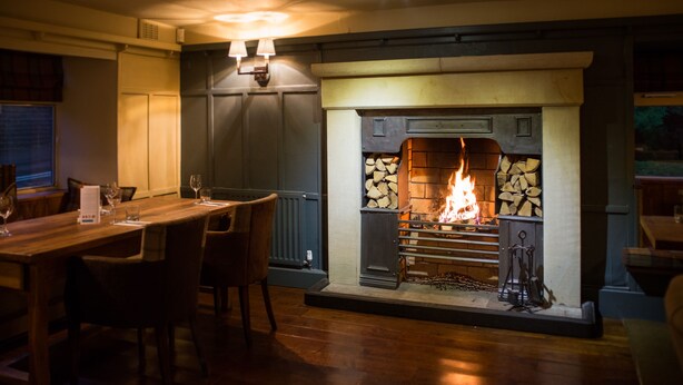 A cozy dining area with a wooden table and chairs, a fireplace with logs, and a wall-mounted light fixture.