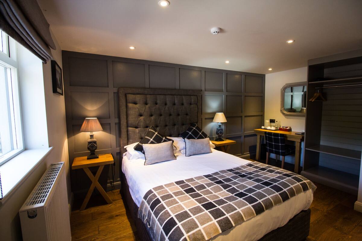 A modern bedroom with a large bed, two bedside tables with lamps, a dresser, and a window with blinds.