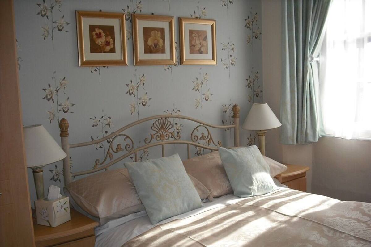 A bedroom with a bed, two bedside tables, a lamp, a nightstand, and a window with curtains.