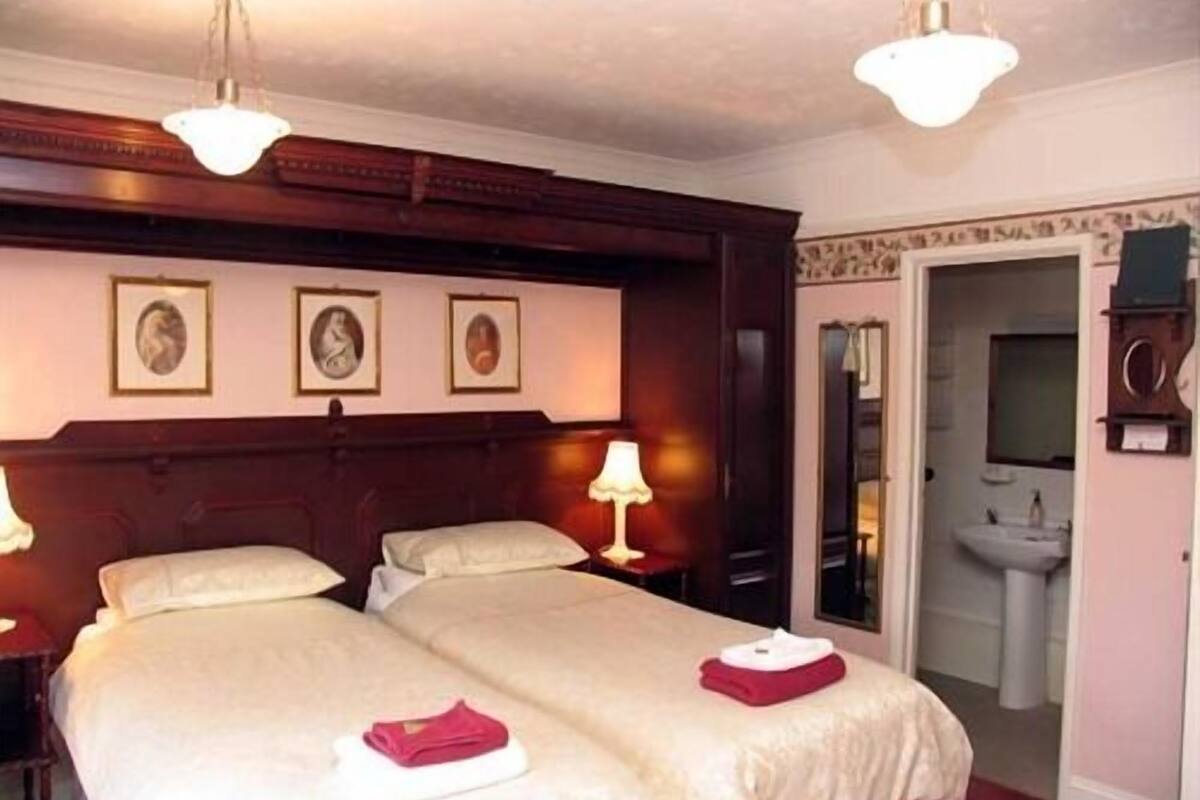 A hotel room with two beds, a nightstand, a lamp, and a door leading to a bathroom.