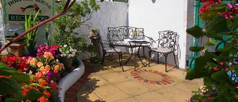 Un patio con mesa y sillas, rodeado de plantas y flores en macetas. Se ve un letrero en el fondo que dice "Dolphin House Reception".