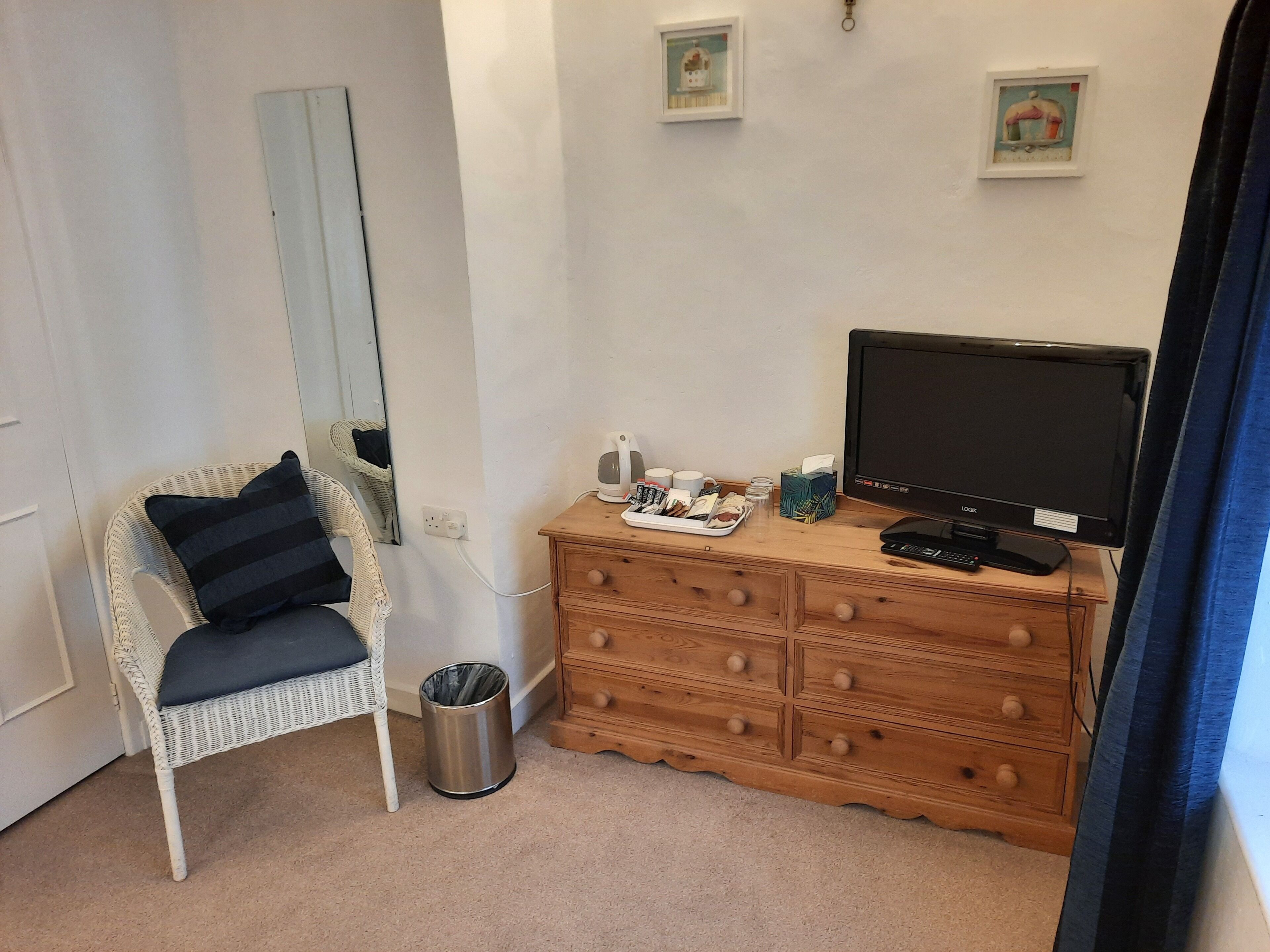 Een slaapkamer met een witte rieten stoel, een houten dressoir, een televisie en een afvalbak.