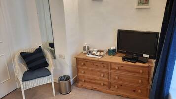 Een slaapkamer met een witte rieten stoel, een houten dressoir, een televisie en een afvalbak.