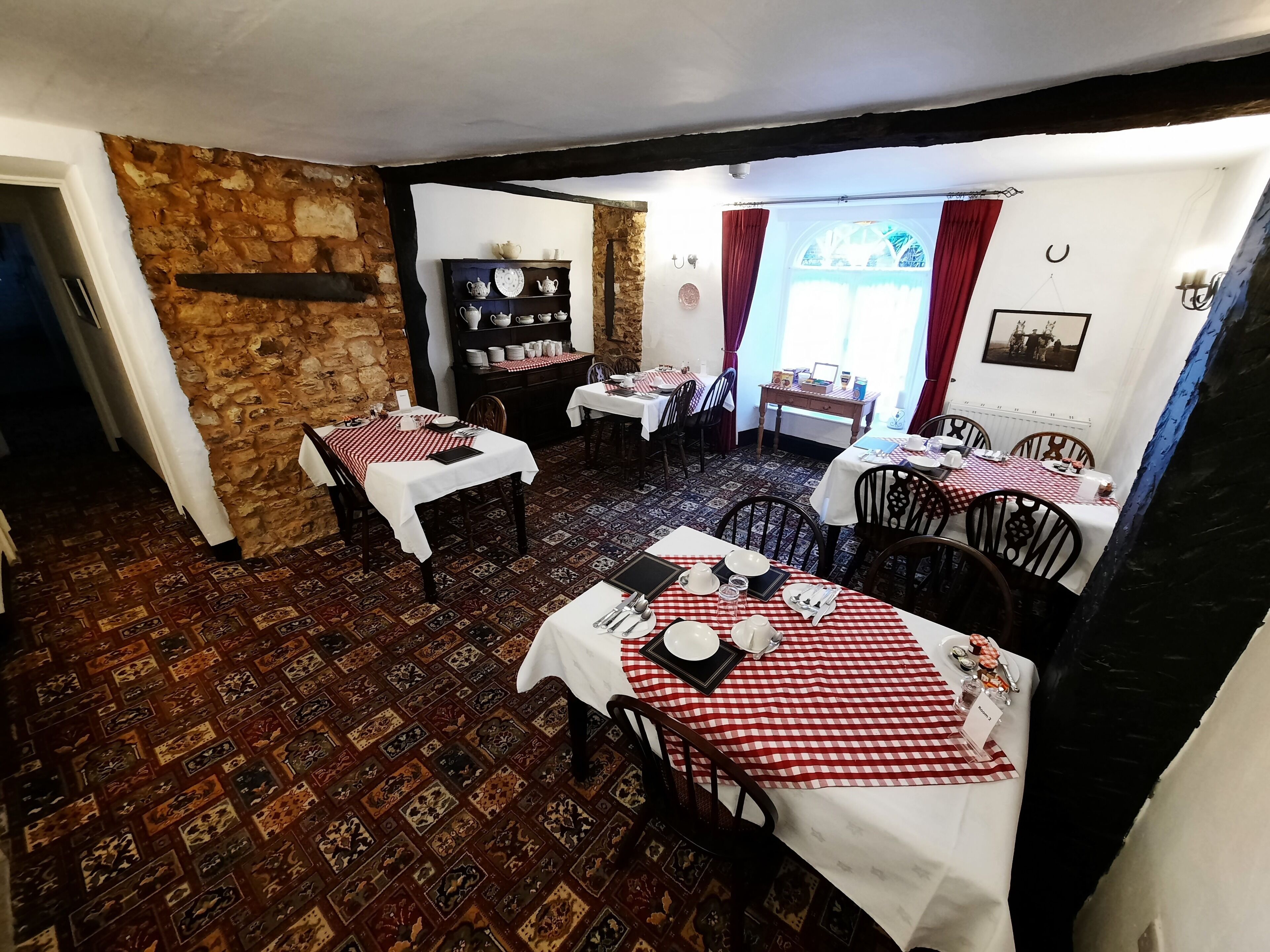 Un comedor con manteles a cuadros, sillas de madera y una pared de piedra. Las mesas están dispuestas con platos, vasos y cubiertos.