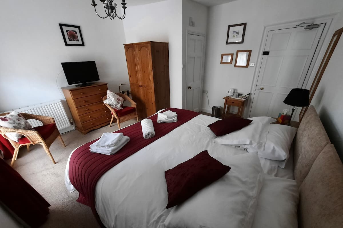 Hotellihuone, jossa on kaksi sänkyä, puinen vaatekaappi, televisio, pieni pöytä lampun kanssa ja tuoli, jossa on punainen koristetyyny.