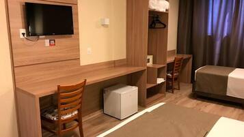 Habitación de hotel con cama, escritorio con silla, televisor, armario y refrigerador pequeño.