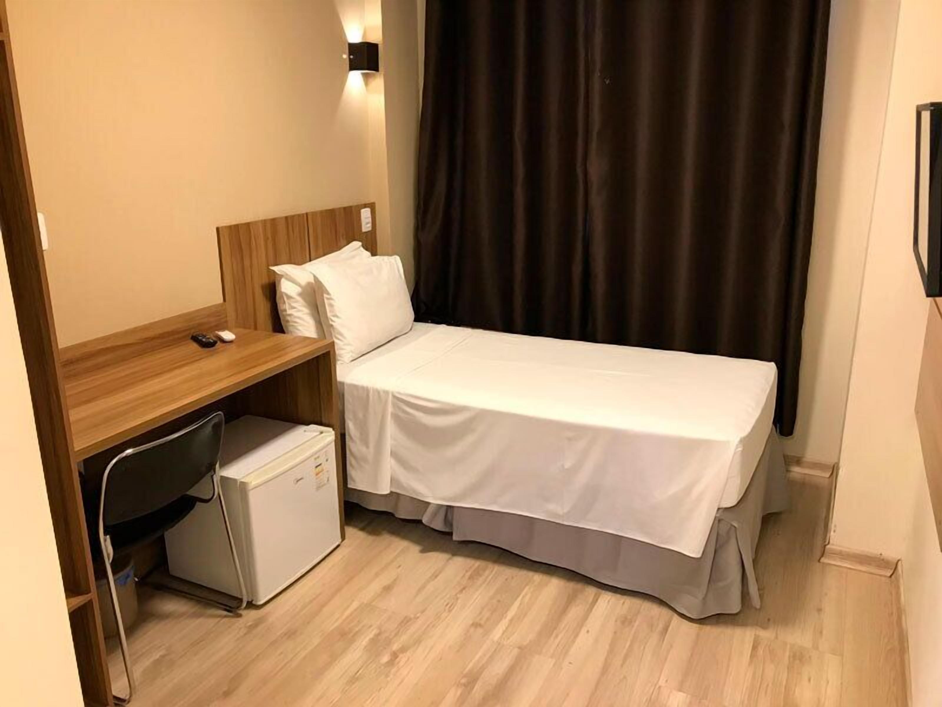 Habitación de hotel compacta con una cama individual, un escritorio pequeño, una silla, un refrigerador y una lámpara fijada a la pared.