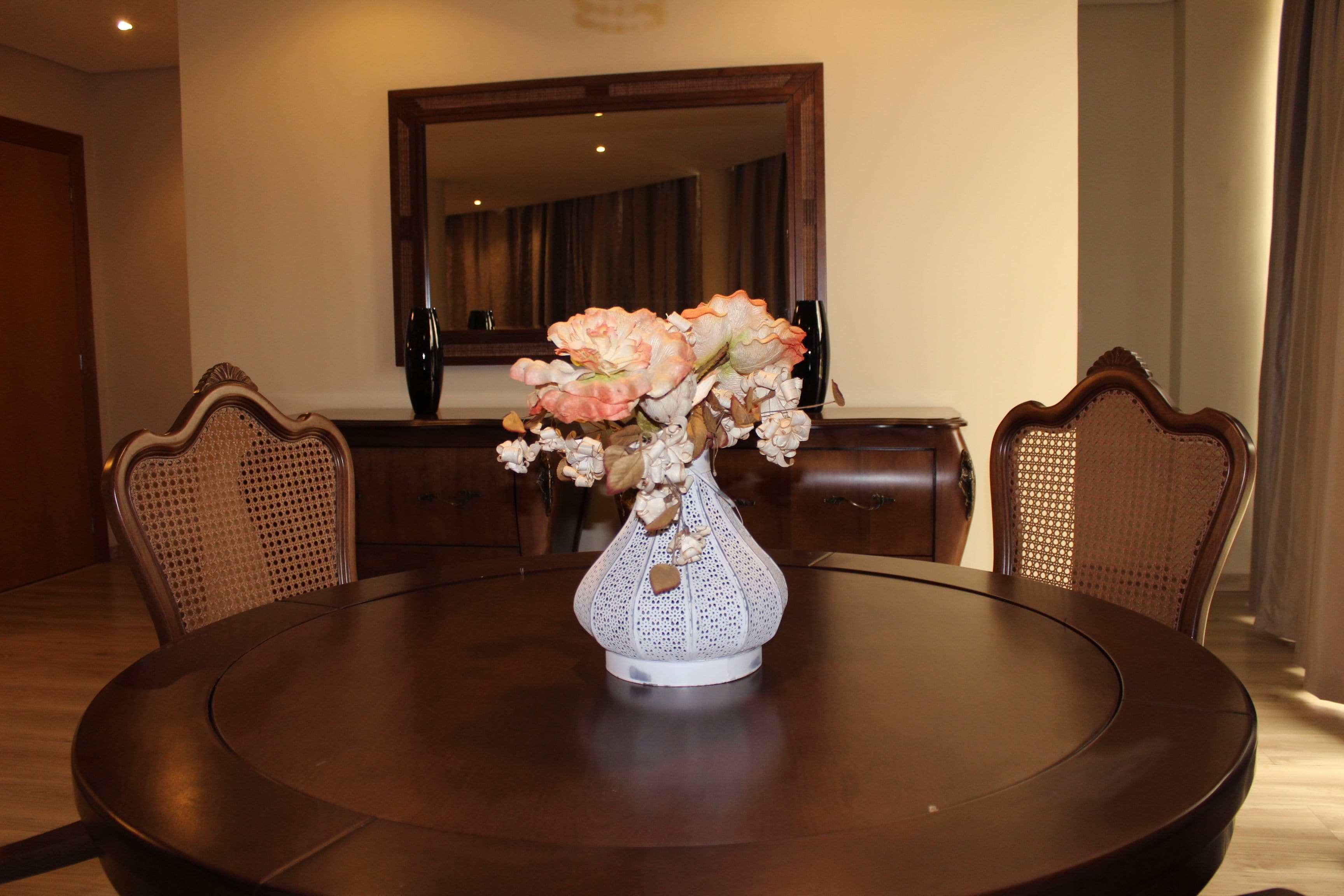 Una mesa redonda de comedor con un jarrón blanco lleno de flores, rodeada por cuatro sillas de madera con respaldo trenzado.