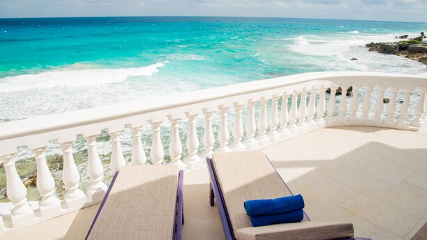 Uma varanda com duas cadeiras de descanso, uma delas com uma toalha azul, com vista para um mar turquesa e ondas quebrando em uma costa rochosa.