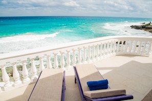 A balcony with two lounge chairs, one with a blue towel, overlooking a turquoise sea with waves crashing onto a rocky shore. - Villa Elena Isla Mujeres (Isla Mujeres)