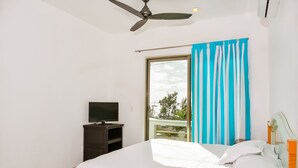Quarto com cama, televisão sobre suporte, ventoinha no teto e janela com cortinas.