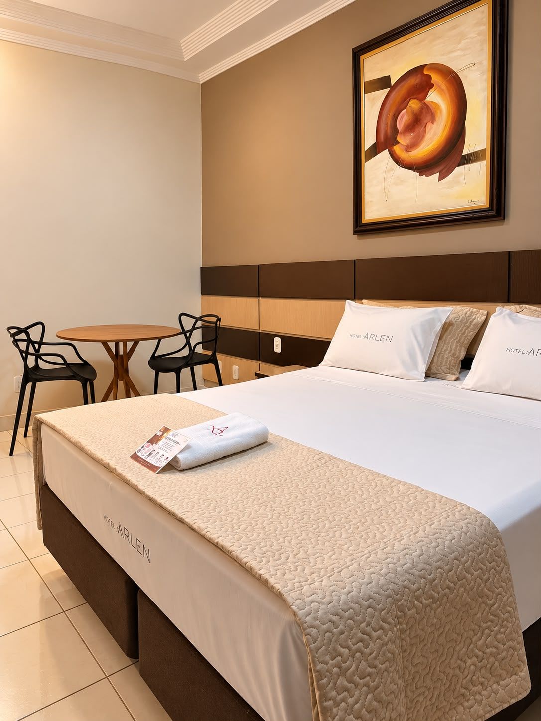 A hotel room with a large bed, a small round table, two chairs, and a painting on the wall.