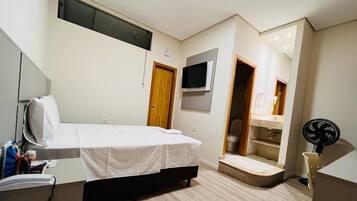 A hotel room with a bed, a TV mounted on the wall, a small desk with a chair, a fan, and a bathroom visible through an open door.