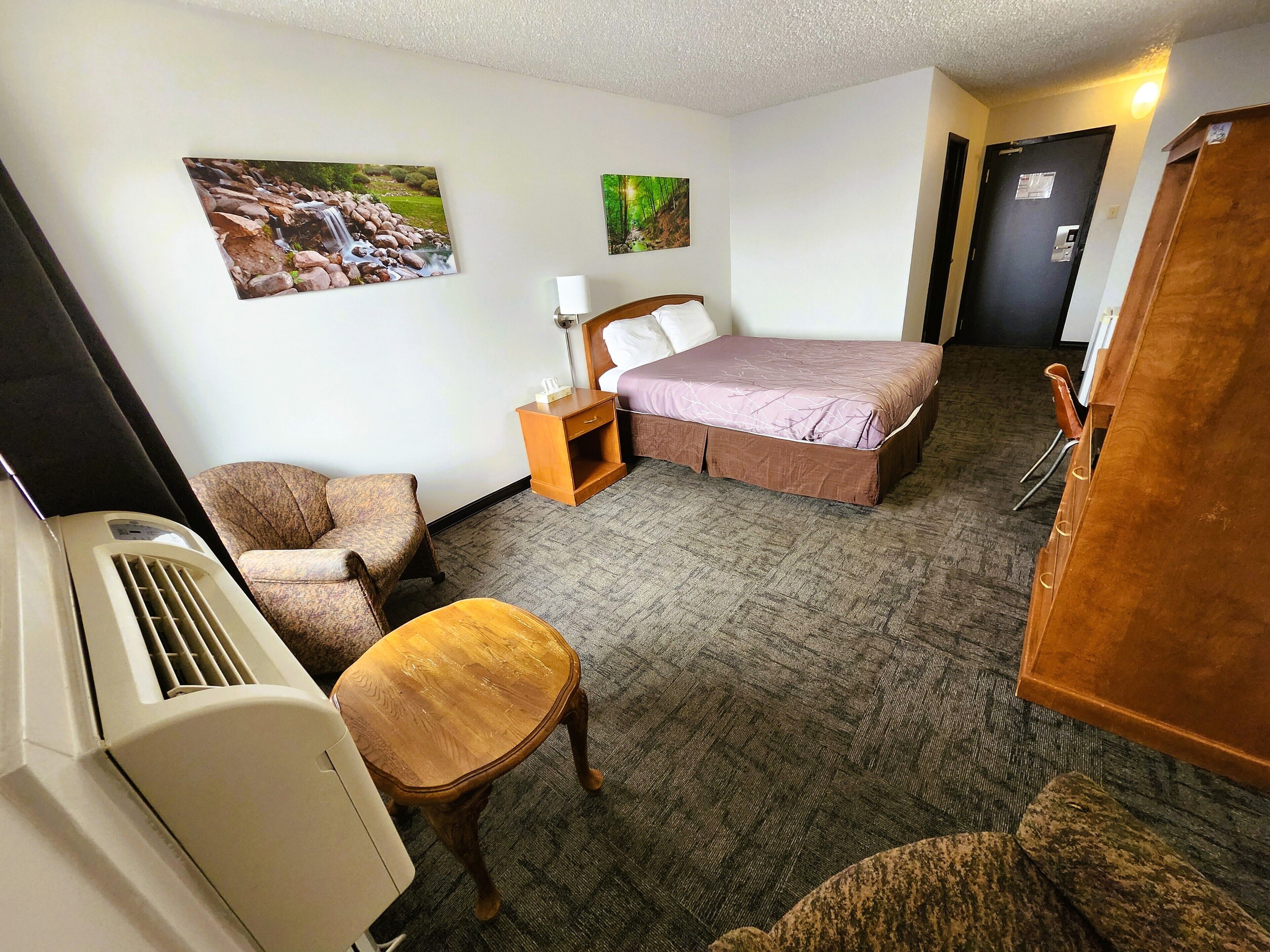 A hotel room with a bed, a chair, a small table, a nightstand, a lamp, and two wall-mounted pictures.