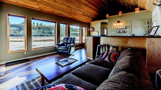 A spacious living room with a wooden ceiling, large windows offering a view of a rocky landscape, a leather sofa, and a coffee table. - Yellowstone River Lodge (Columbus)