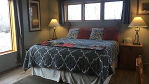 A bedroom with a bed, two bedside lamps, a window with curtains, a framed picture on the wall, and a wooden chair. - Yellowstone River Lodge (Columbus)