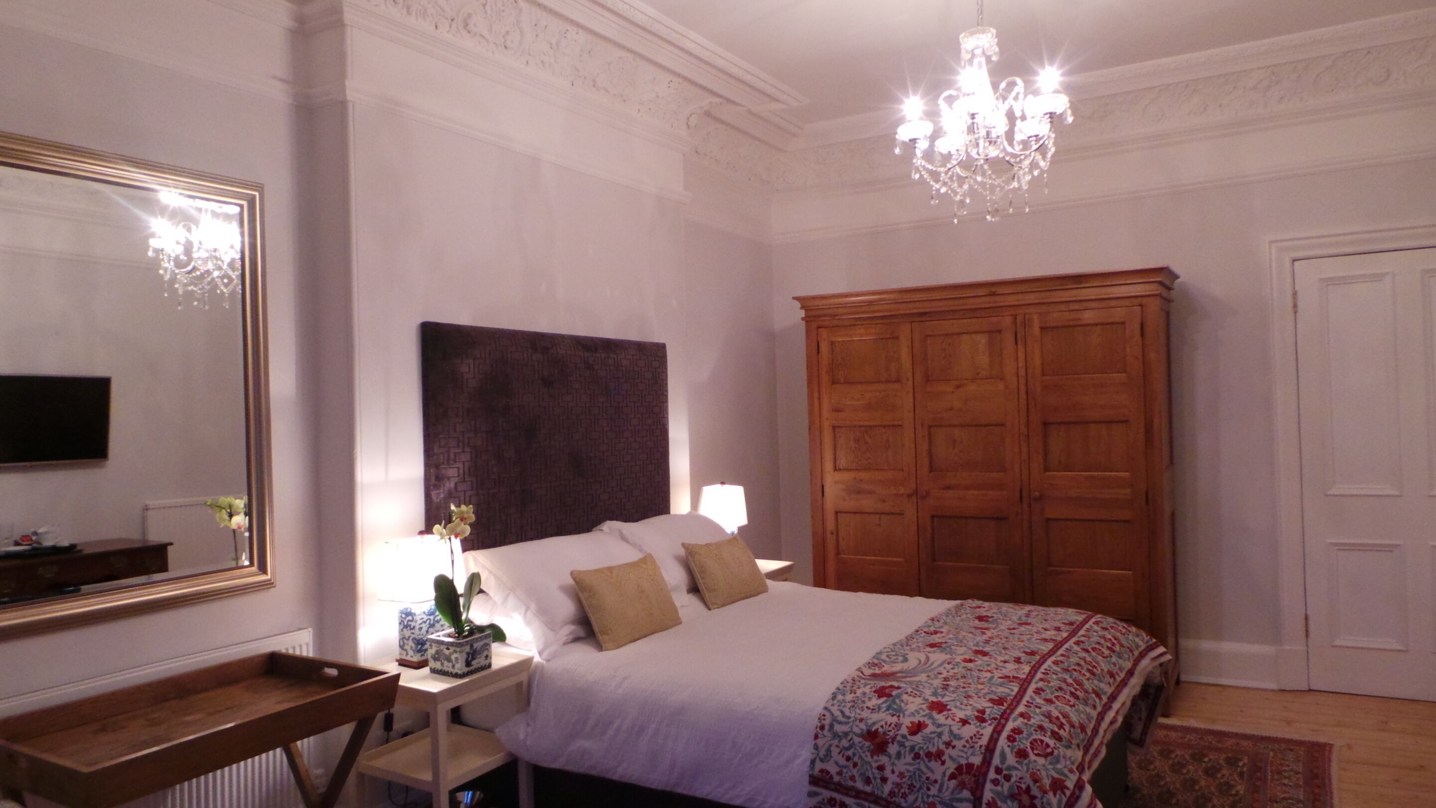 A bedroom with a large bed, a wooden wardrobe, a chandelier, a mirror, and a bedside table with a lamp and a plant.