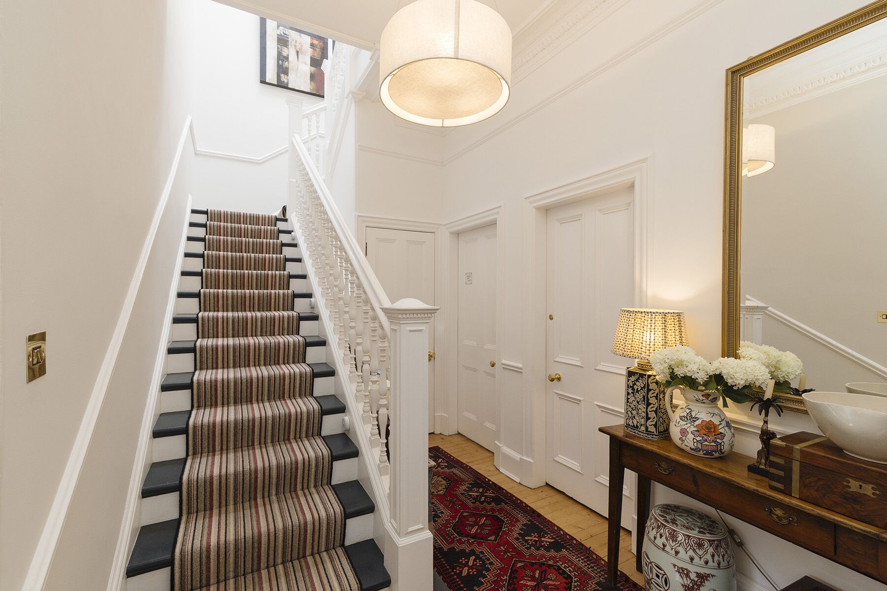 A staircase with striped carpet, a chandelier, a mirror, and a console table with decorative items.