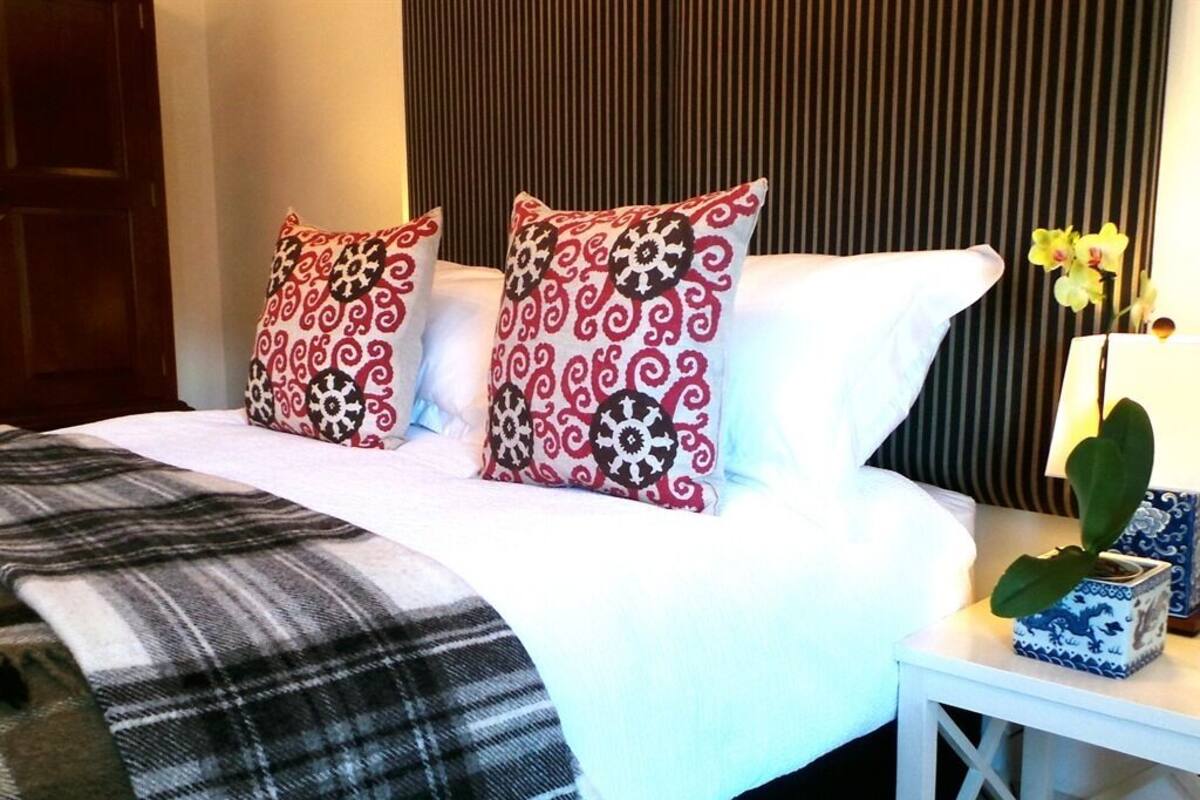 A neatly made bed with patterned pillows, a plaid blanket, and a small table with a vase of flowers.