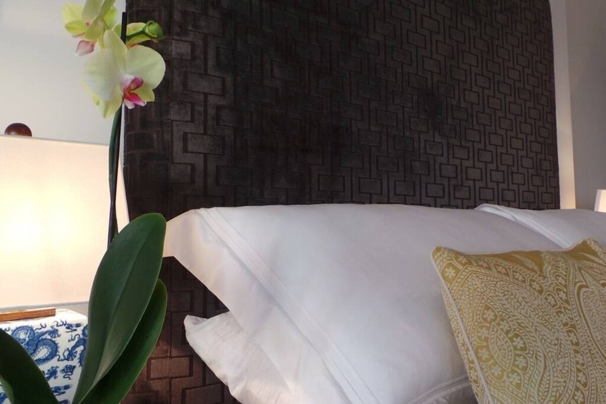 A neatly made bed with white linens and a decorative pillow, a potted orchid plant, and a patterned wall behind the bed.