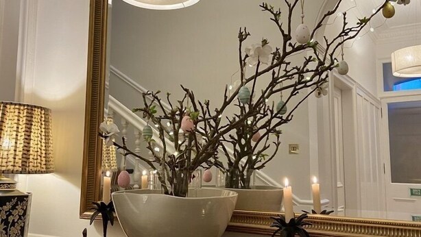 Une pièce avec un grand miroir, une commode en bois, un vase blanc orné de branches et un luminaire suspendu.