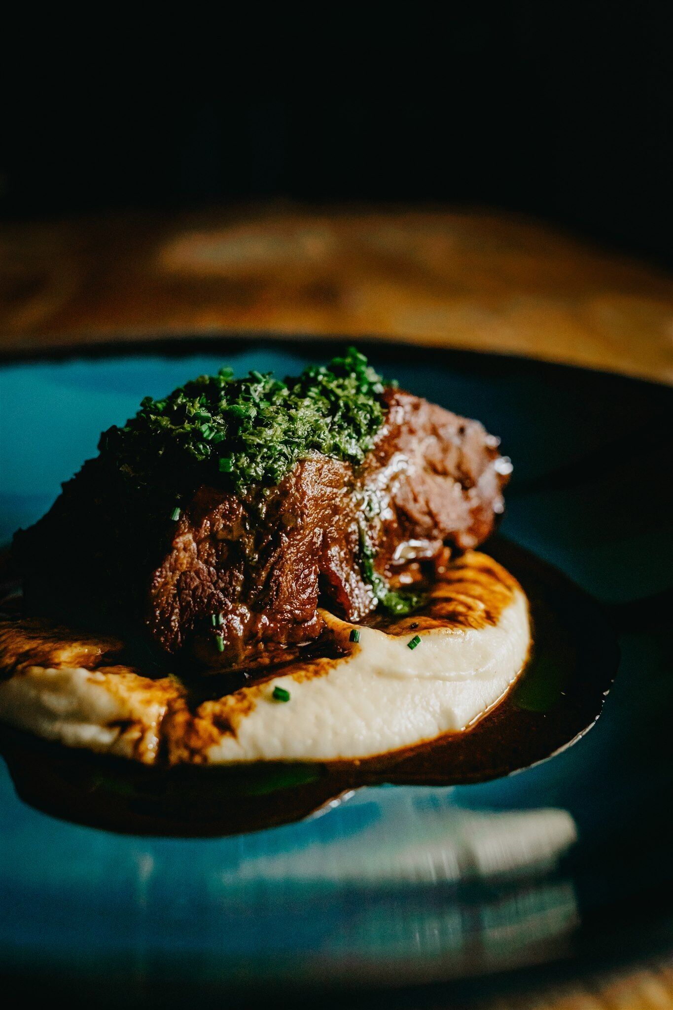 A close-up of a dish with meat and a creamy base, garnished with green herbs, served on a dark plate.