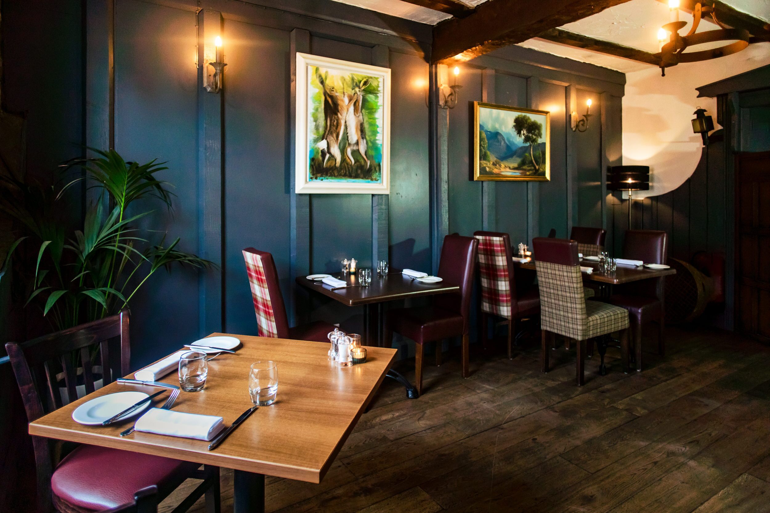 A restaurant interior with wooden tables and chairs, framed artwork on the walls, and a potted plant.