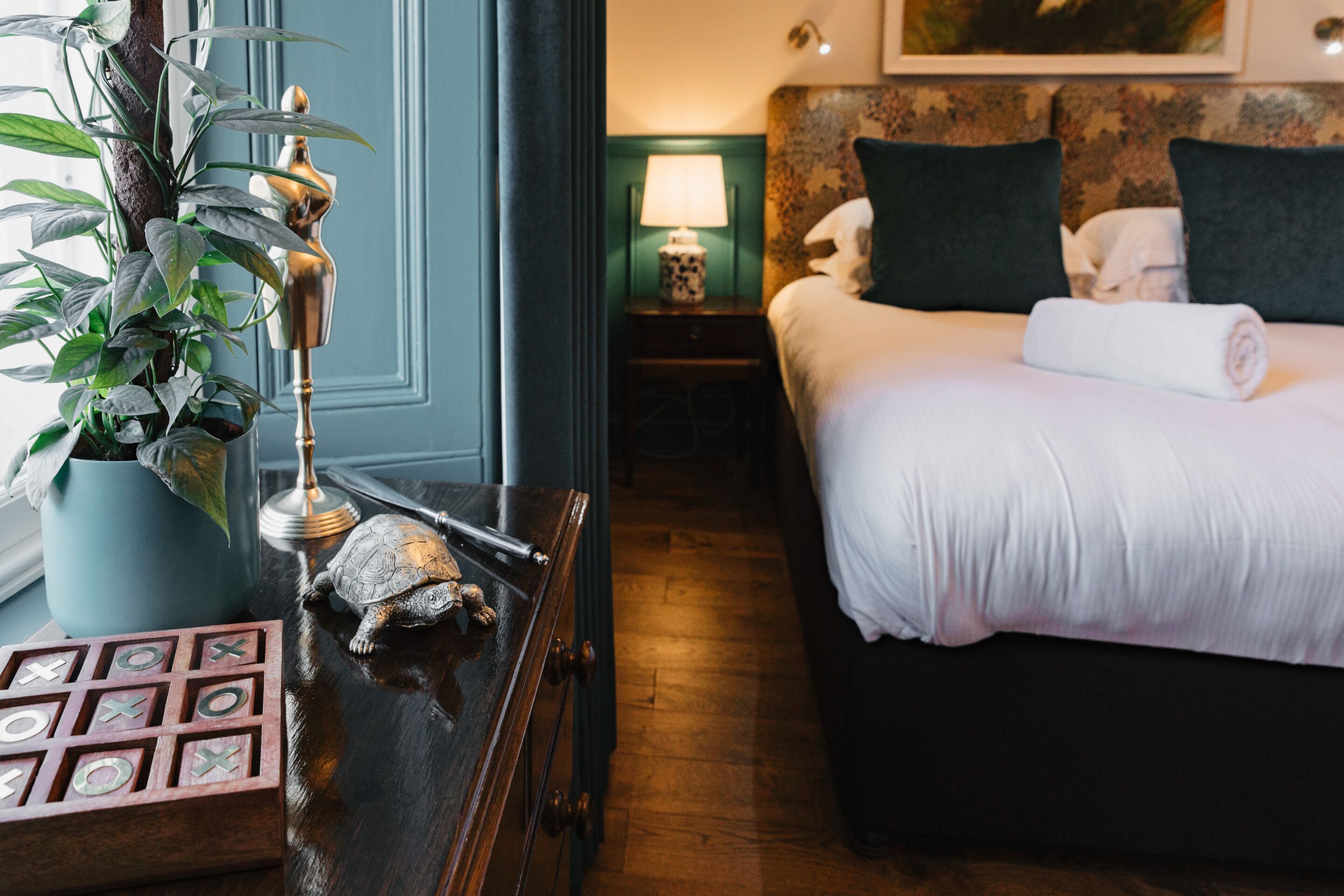 A hotel room with a bed, a nightstand with a lamp, a potted plant, and a chessboard.