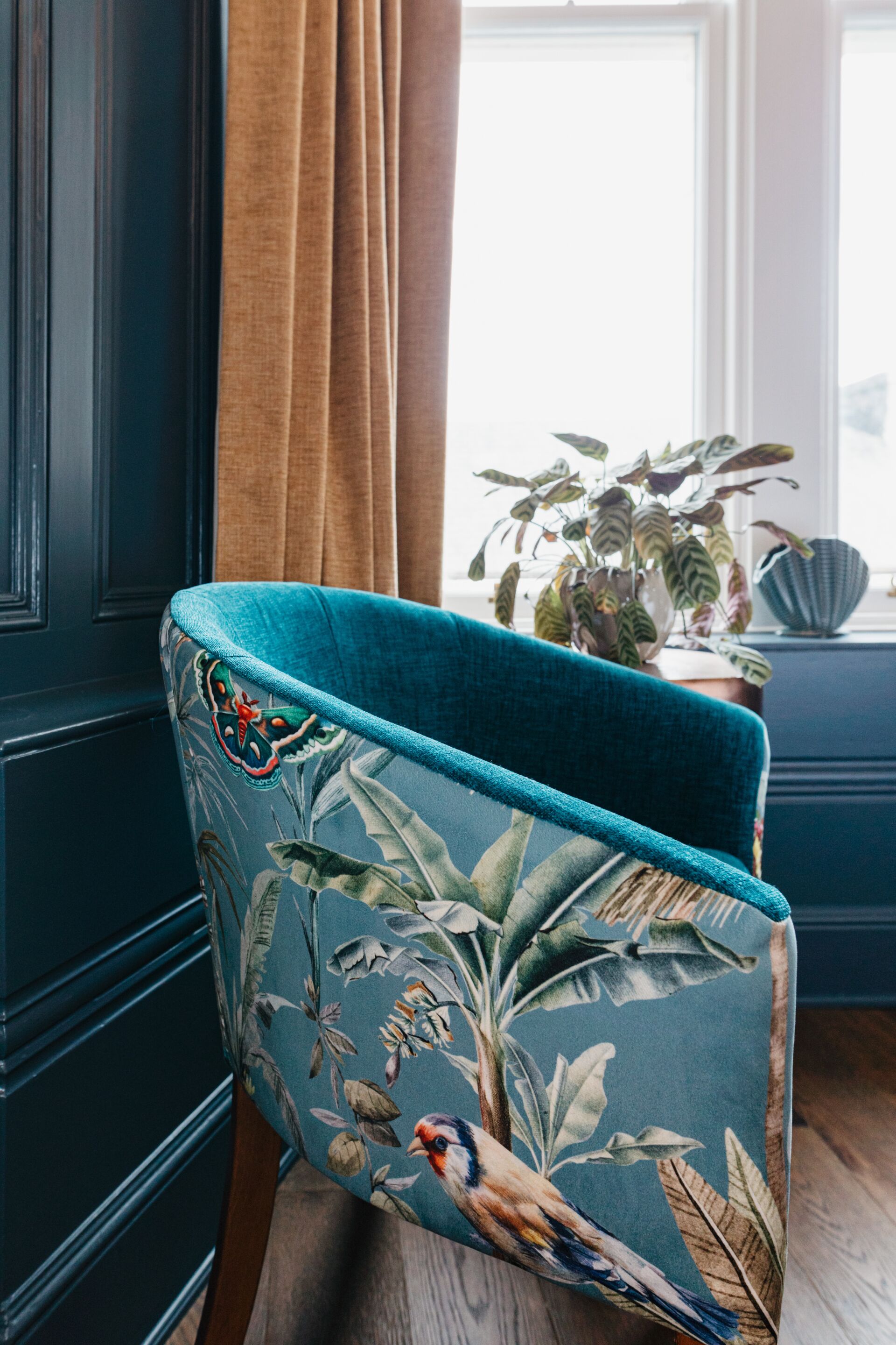 A chair with a floral pattern and a blue cushion, positioned in front of a window with curtains.