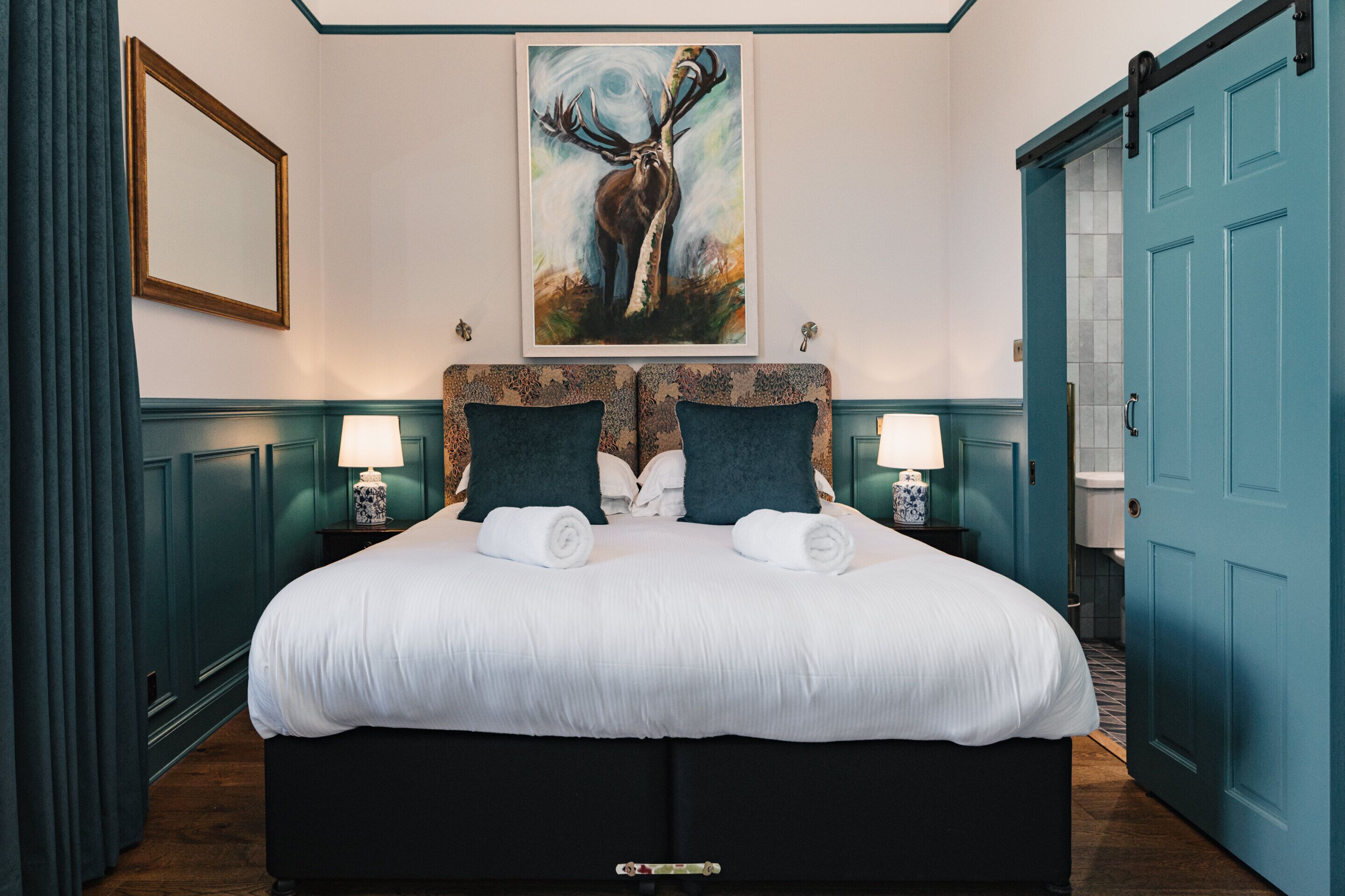 A large bed with a white duvet, two dark pillows, and a headboard with a floral pattern. There is a painting of a deer on the wall above the bed.