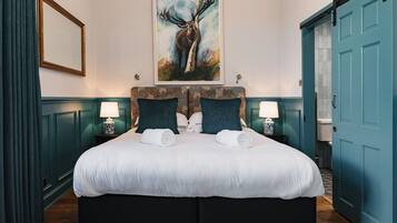 A large bed with a white duvet, two dark pillows, and a headboard with a floral pattern. There is a painting of a deer on the wall above the bed.