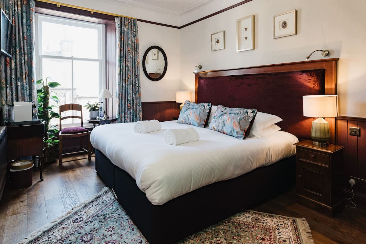 A hotel room with a large bed, two bedside tables, a desk, a chair, a potted plant, and a window with curtains.