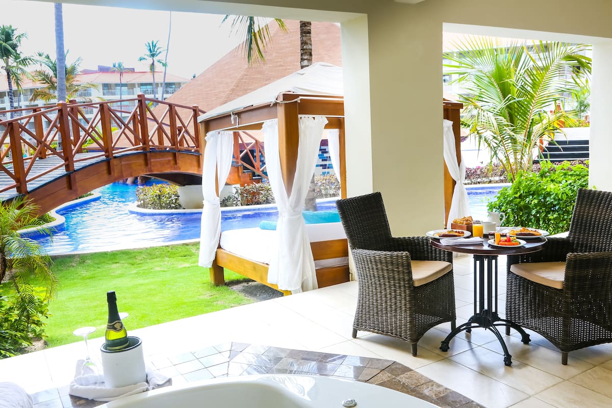A balcony with a view of a swimming pool, a bed with a canopy, wicker chairs, and a table with breakfast.