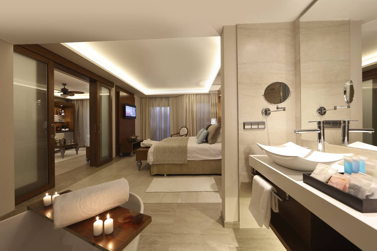 A modern hotel room with a large bed, a sitting area with candles, a vanity with a mirror, and a television in the background.
