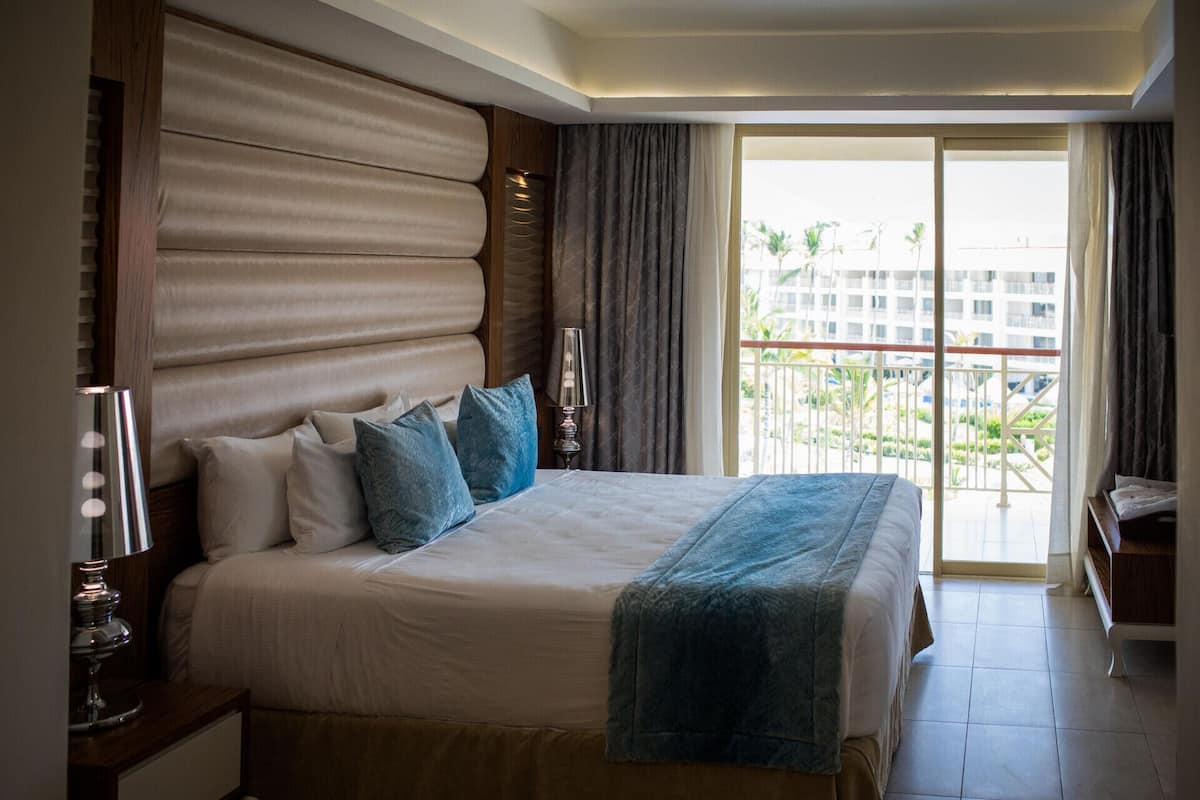 A hotel room with a large bed, a bedside table, a lamp, and a view of a building and greenery through the window.